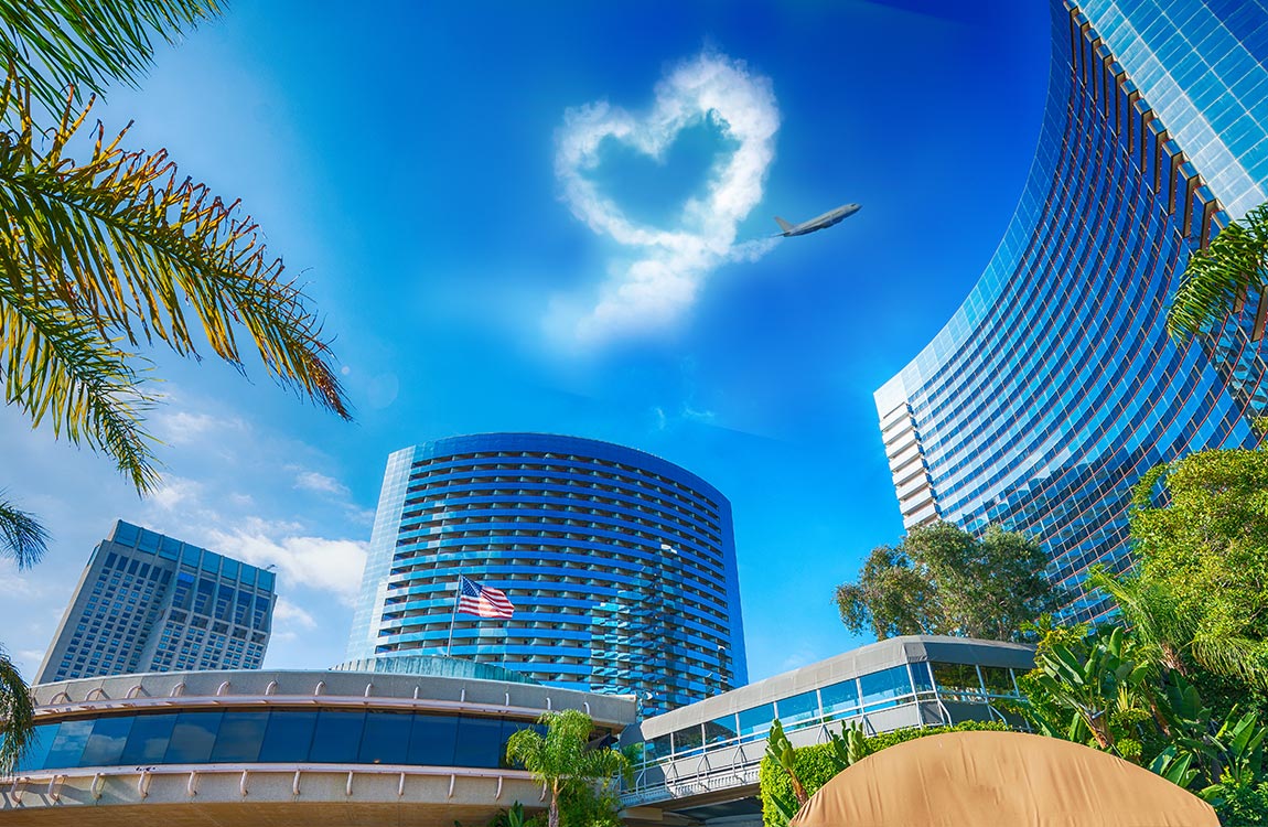 The Marriott Marquis San Diego Marina and other buildings surrounded by palm trees, with a bright blue sky containing a commercial airlines with a heart shaped contrail behind it 