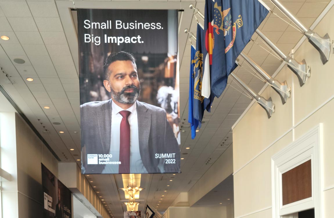 A banner hanging from the ceiling featuring Aventure president Talha Faruqi and the words "Small Business Big Impact" on display next to U.S. flags, in a long cooridor 