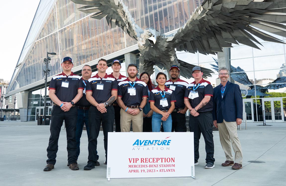 Ten people stand in front of a huge falcon statue in front of a sports stadium, with a sign saying "Aventure Aiation VIP Reception, Mercedes-Benz Stadium April 19, Atlanta" 