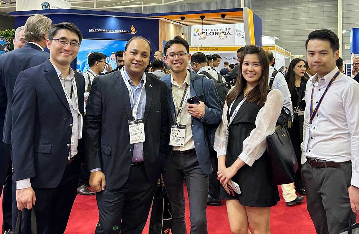 Five conference attendees smile towards camera on the trade show floor, with booths, displays, and a crowd of people behind them 