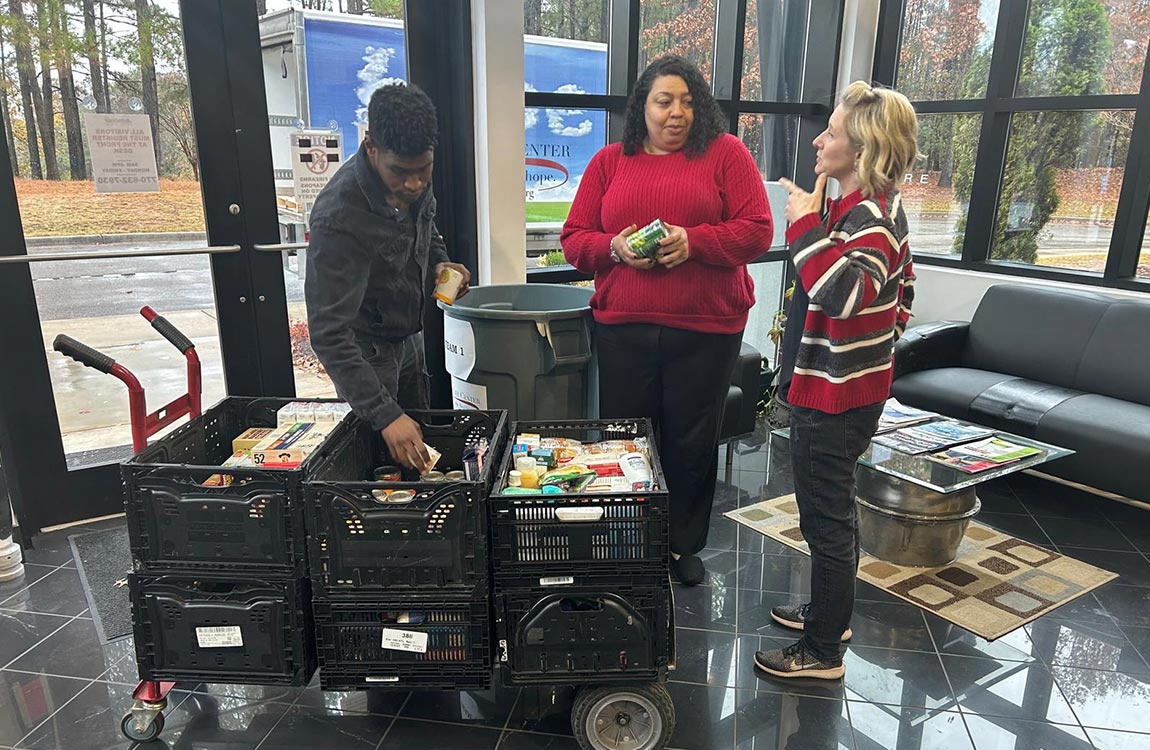 A man and two women load a card full of canned food in a large lobby, with a truck visible outside through large glass windows 