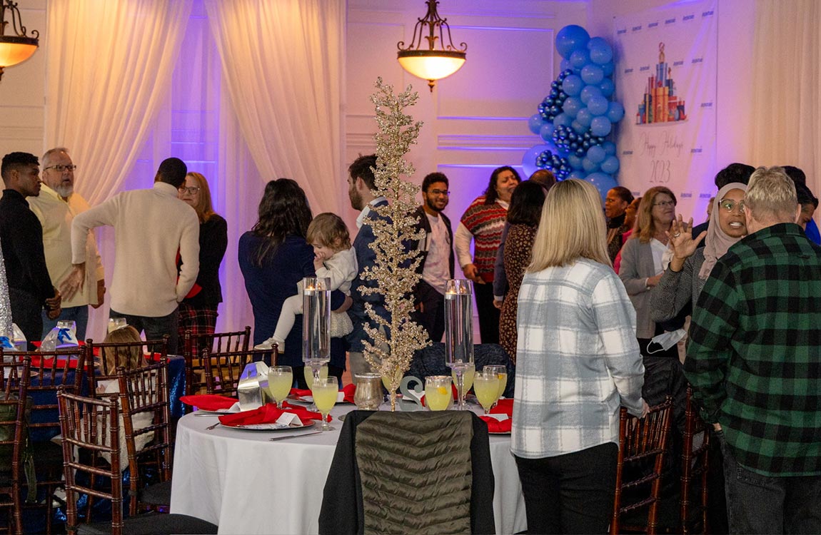 Almost two dozen people socialize in a ballroom full of dining tables and holiday decorations 