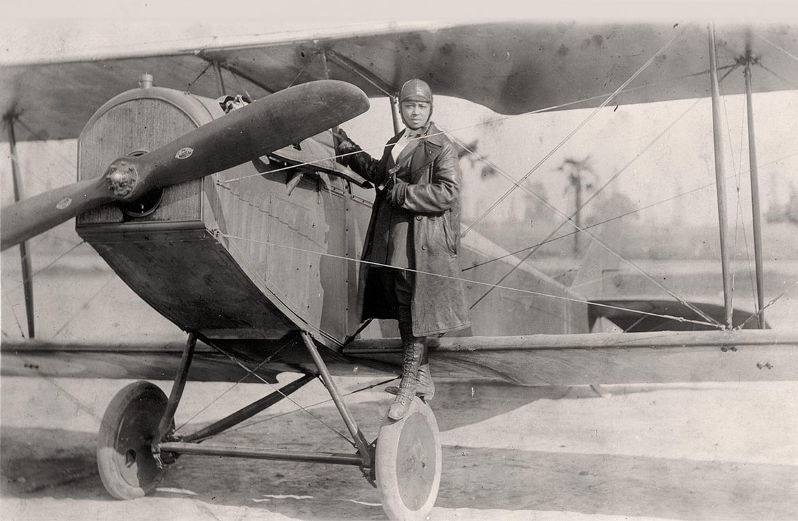 A female African American aviator (Bessie Coleman) in a long trenchcoat posed for a photo before climbing into the cockpit of a 1920s biplane 