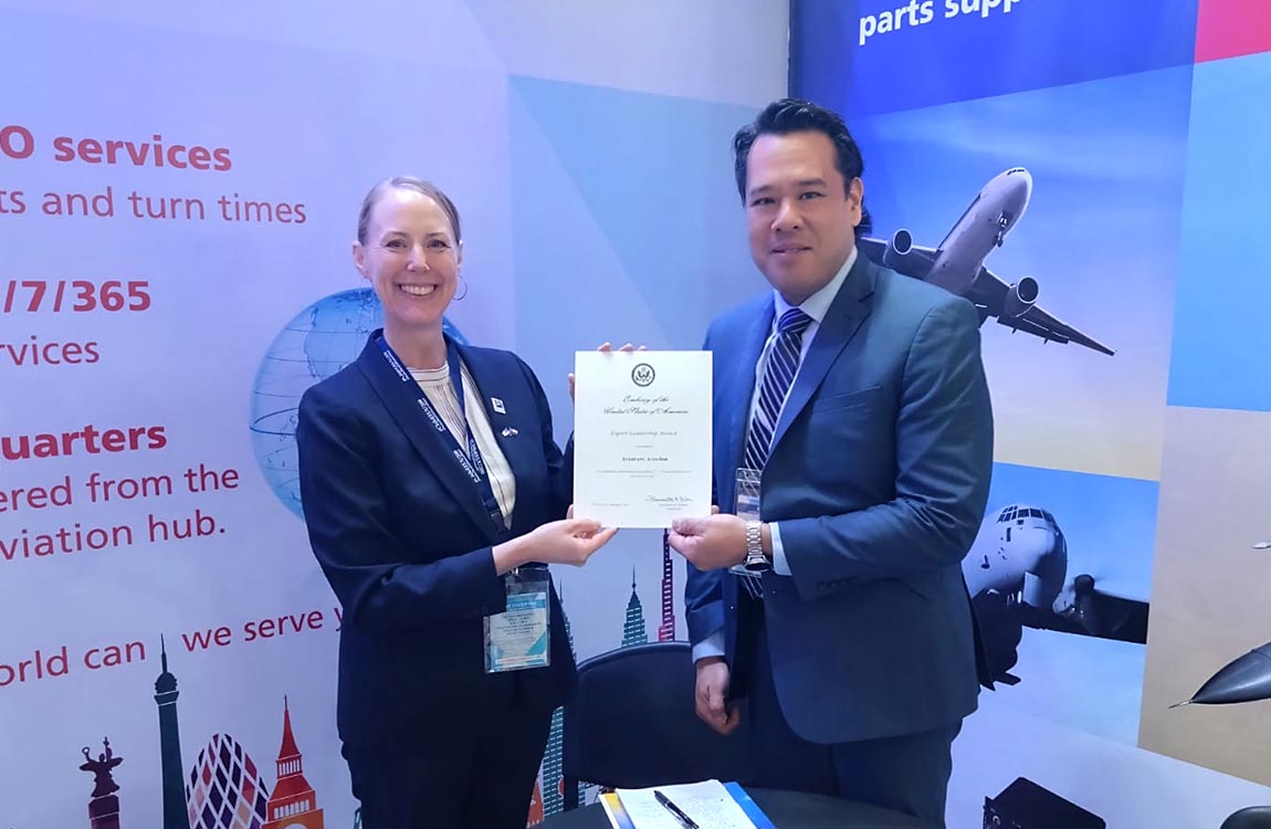 A smiling woman and man in business attire hold up an award in a tradeshow booth with photos of airplanes in the background 