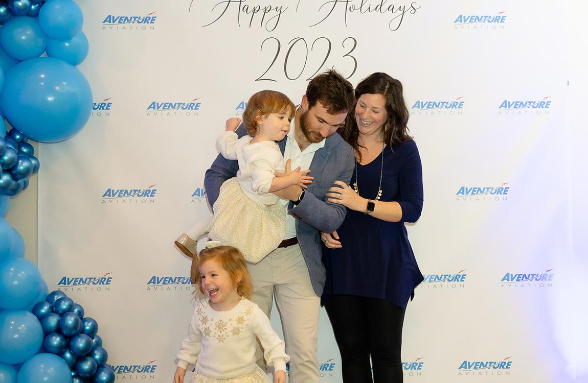 A family of four smiling in front of a "step and repeat" backdrop saying "Aventure Aviation Happy Holidays 2023 " with blue balloons 