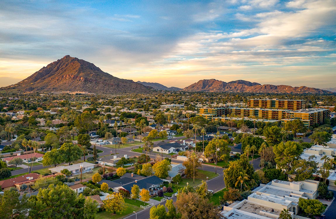 Low-rise buildings surrounded by trees with mountains in the background under a bright, cloudy sky 