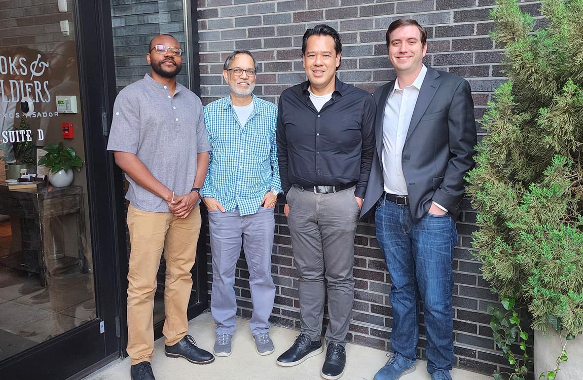Four men, dressed in business casual, smile towards the camera while outside a place of business 