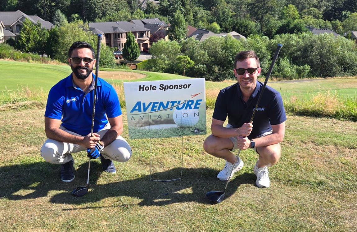 Two men with golf clubs on a golf green kneel by a sign that says "Hole Sponsor Aventure Aviation" 