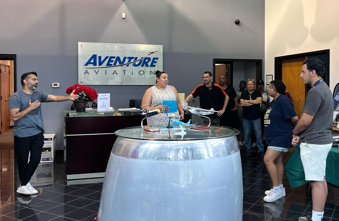 Eight people interacting in a bulding lobby, with a sign on the wall saying "Aventure Aviation" and model aircraft on a table made of an aircraft engine 