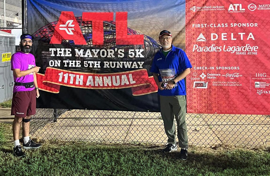 Two men dressed in running clothes standing outside, next to a sign that says "ATL - The Mayor's 5K on the 5th Runway 11th Annual 