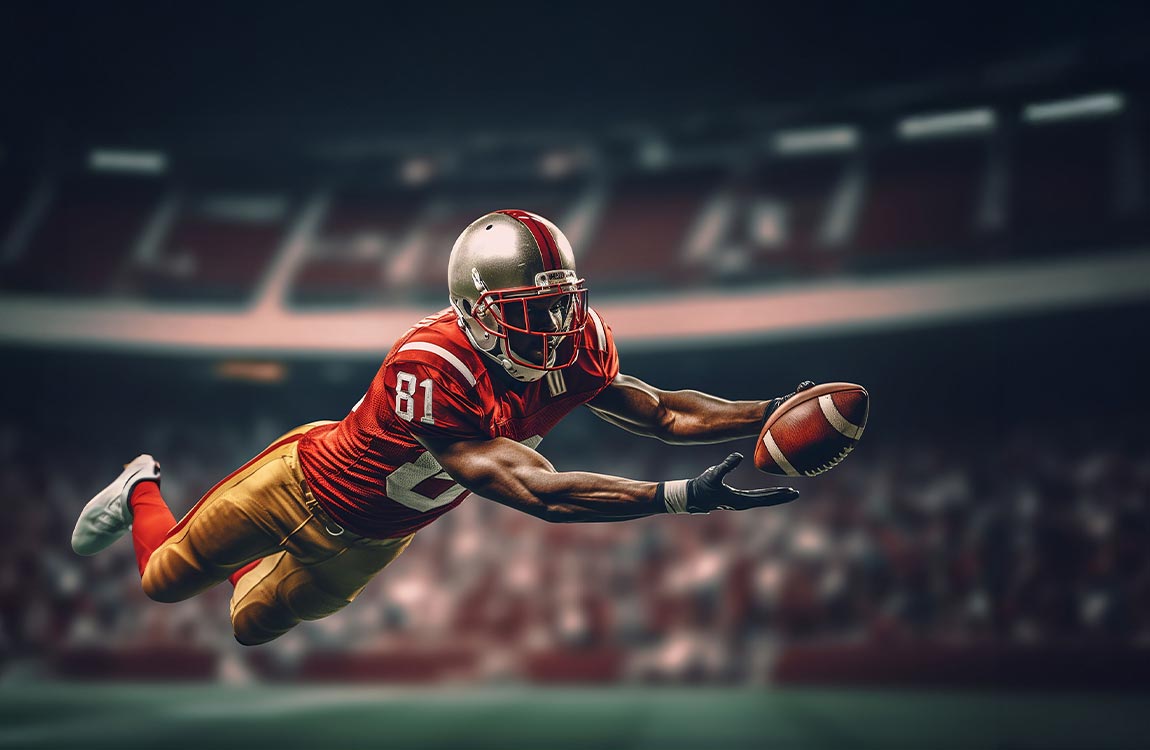 An American football player dives through the air to catch a football inside a football stadium during a night game  