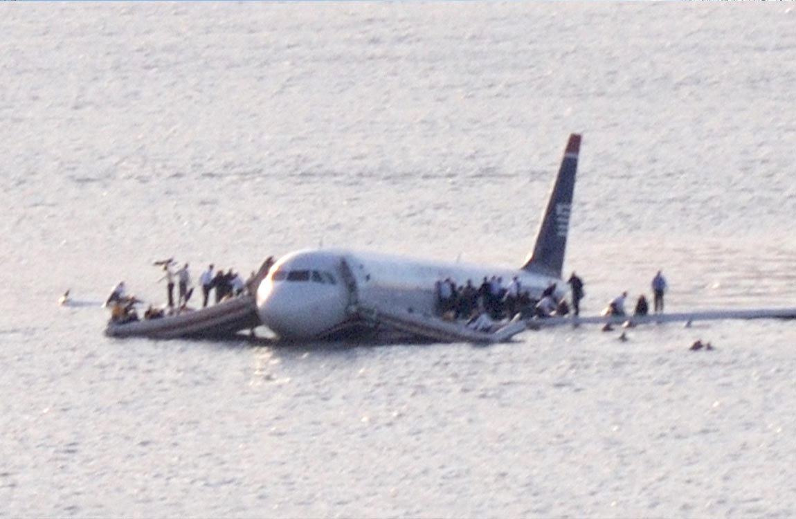 A commercial aircraft floats on a body of water, with people standing on its wings and evacuation slides 