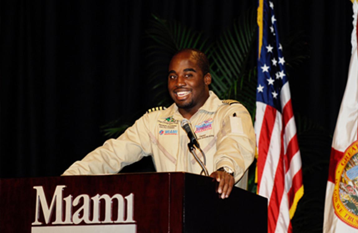 A Black man in a pilot's uniform at a podium saying "Miami" in front of an American flag 