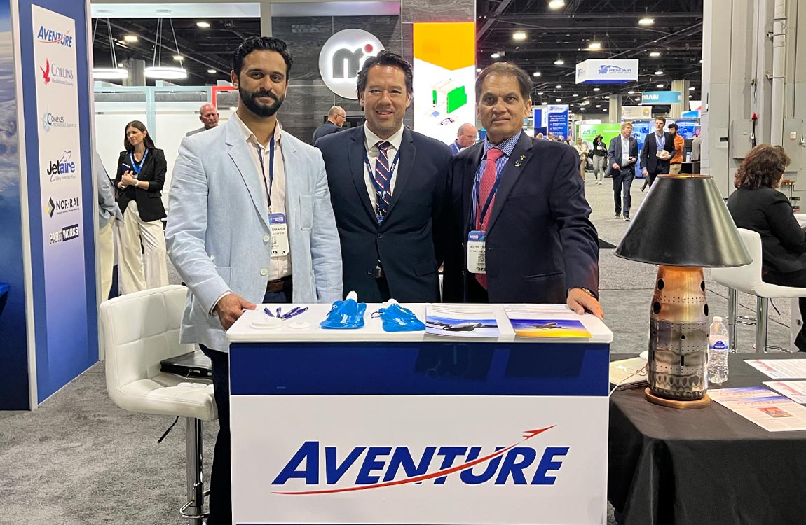 Three men in business suites standing above a counter saying "Aventure" on the floor of a large tradeshow 