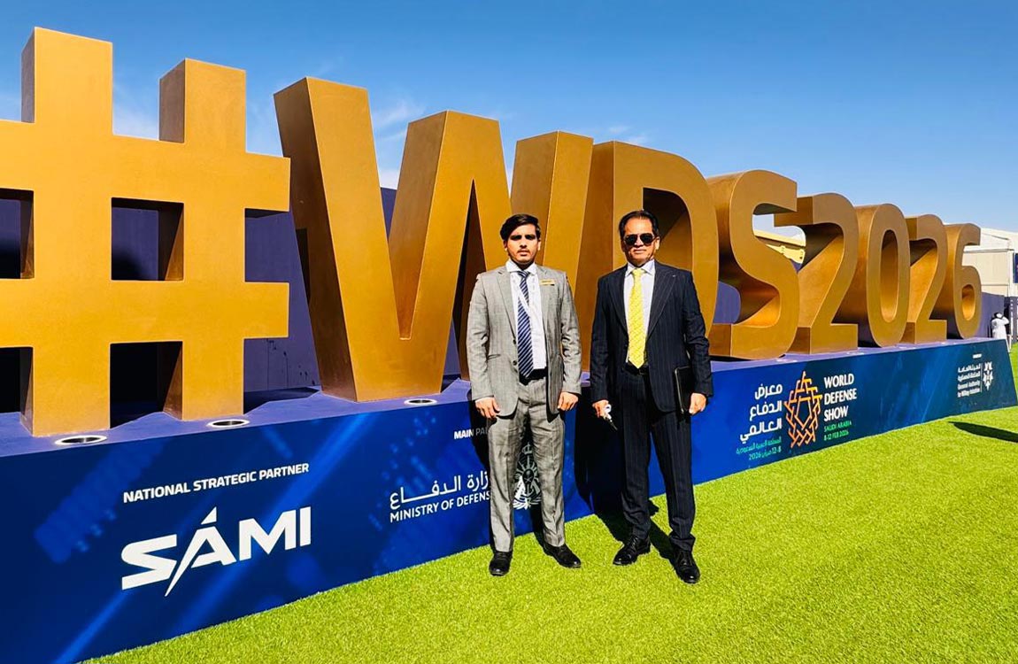 Two men in business suits stand outside by a huge display saying #WDS2026 under a blue sky 
