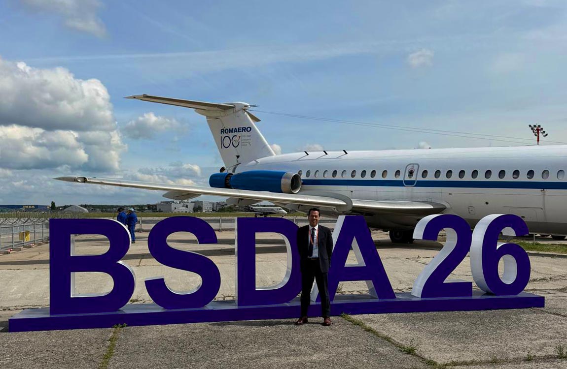 Man wearing a suit standing in front of huge letters saying BSDA26 on an airport tarmac, with a large commercial jet in the background under  blue sky 