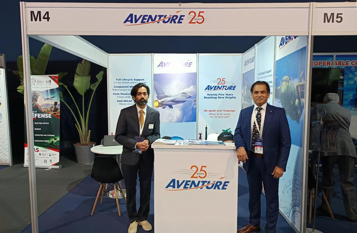 Five people stand inside a large tradeshow booth that says "Aventure 25 years" and has multiple color photos of aircraft, and a table with seats 