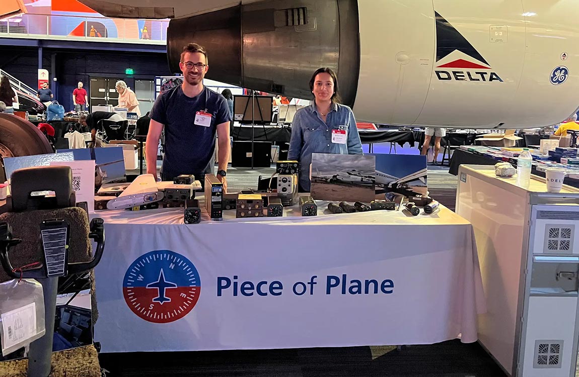 A man and a woman stand behind a table that says "Peace of Plane" with the company logo, in a large room containing aviation parts and a full size Delta Airlines jet 